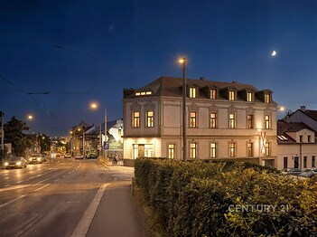 Historická budova v nočním osvětlení, nacházející se na rohu ulice. Budova má klasickou fasádu a velká osvětlená okna.