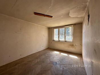 Prázdná místnost s parketovou podlahou a velkými okny, vyžadující rekonstrukci, s popraskanou stěnou a stropem.