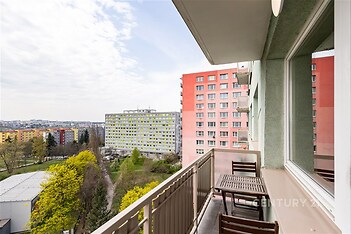 Balkon s výhledem na město, dřevěný nábytek, byt v panelovém domě, světlá fasáda a okolní zeleň vytváří příjemnou atmosféru pro relaxaci.