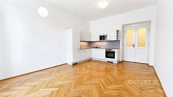 Světlá kuchyň s parketovou podlahou a moderními spotřebiči, světlé stěny a velká okna zajišťují dostatek přirozeného světla.