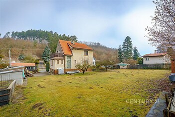 Prostorná zahrada s rodinným domem, doplněná zeleným trávníkem a vzrostlými stromy, nabízí klidné prostředí a soukromí pro bydlení.