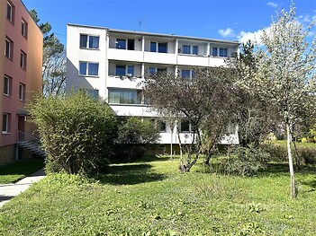 Čtyřpodlažní bytový dům s bílou fasádou, obklopený zeleným trávníkem a stromy, umístěný na klidném místě v rezidenční čtvrti.