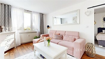 Světlý obývací pokoj s parketovou podlahou, velká okna a moderní design. Přirozené osvětlení a stylové dekorace vytvářejí útulnou atmosféru.