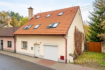 Rodinný dům s červenou střešní krytinou a světlou fasádou, doplněný garážovými vraty a střešními okny, situován na klidné ulici poblíž zeleně.