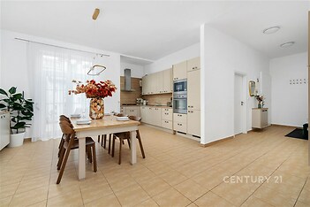 Prostorná kuchyň s dlažbou, moderními spotřebiči a světlým nábytkem, jídelní stůl s židlemi, velká okna poskytující dostatek přirozeného světla