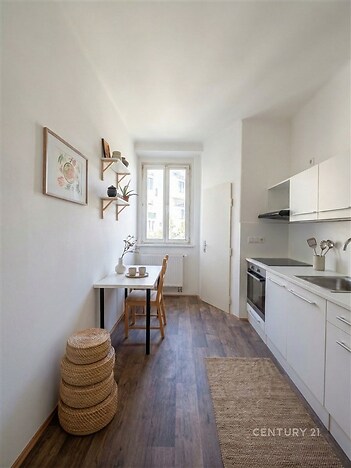 Kompaktní kuchyň s moderními spotřebiči, dřevěnou podlahou a světlými skříňkami, velké okno poskytuje dostatek přirozeného světla.