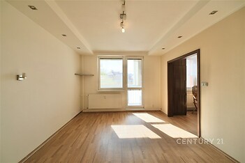 Světlý obývací pokoj s parketovou podlahou, velká okna zajišťují dostatek přirozeného světla, moderní osvětlení a neutrální barvy pro útulný vzhled.