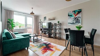 Světlý obývací pokoj s parketovou podlahou, velkými okny a moderním nábytkem, včetně jídelního stolu a pohodlné sedací soupravy.