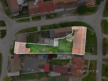 Pohled z ptačí perspektivy na pozemek s dvěma stavbami a zahradou, umístěný v klidné rezidenční oblasti s červenými střechami domů.