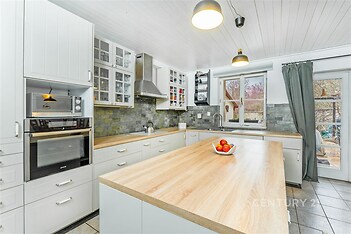 Moderní kuchyň s dřevěnou pracovní deskou, vestavěnými spotřebiči a velkými okny. Světlý prostor je doplněn dlažbou a bílými skříněmi.