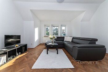 Světlý obývací pokoj s parketovou podlahou, moderní pohovkou a výhledem z velkých oken, vytváří příjemnou atmosféru pro odpočinek.