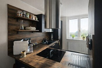 Moderní kuchyň s dřevěnou linkou a velkými okny, která propouštějí dostatek světla, doplněná elegantními černými spotřebiči.