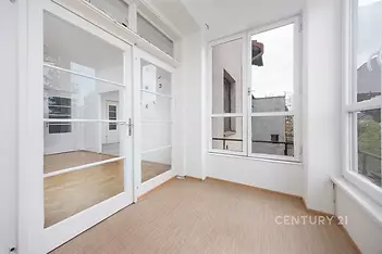 Světlá veranda s velkými okny, parketovou podlahou a moderním designem, nabízející výhled do klidného okolí.
