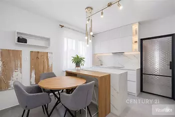Moderní kuchyň s mramorovou pracovní deskou, vestavěnými skříněmi a stylovým osvětlením. Světlý interiér s dřevěným kulatým jídelním stolem.