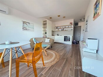 Světlý obývací pokoj s dřevěnou podlahou, moderní kuchyňskou linkou a velkými okny. Skandinávský styl a klimatizace zajišťují komfort.