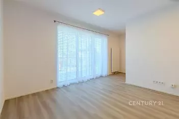 Světlý obývací pokoj s parketovou podlahou, velká okna s bílými závěsy, moderní design a prostor pro relaxaci.