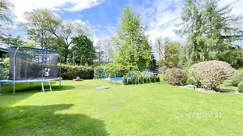 Zahrada s trampolínou a bazénem, udržovaný trávník, obklopená vzrostlou zelení a stromy, ideální pro rodinné aktivity či relaxaci.
