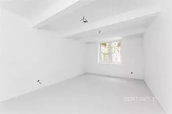 Prázdná místnost s bílými stěnami a stropními trámy, velkým oknem a světlou podlahou, připravená k dokončení nebo rekonstrukci.