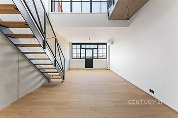 Světlý obývací pokoj s parketovou podlahou a velkými okny, moderní design s kovovým schodištěm a betonovým stropem.