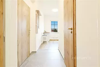 Světlá chodba s keramickou dlažbou, dřevěnými dveřmi a velkým oknem, moderní úprava interiéru, navazuje na obytný prostor.
