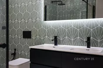 Moderní koupelna s tmavou dlažbou, designové obklady a velké zrcadlo. Elegantní umyvadlo s integrovaným osvětlením a černé doplňky.