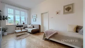 Světlý obývací pokoj s parketovou podlahou, velká okna zajišťují přirozené světlo, moderní dřevěný nábytek dodává elegantní vzhled.
