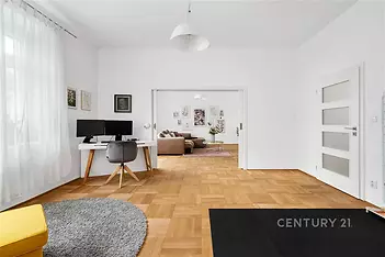 Prostorný obývací pokoj s parketovou podlahou a velkými okny, moderní bílý interiér, pracovní koutek s psacím stolem a pohodlným křeslem.