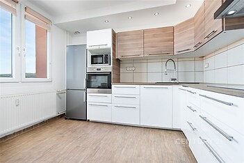 Moderní kuchyň se světlou dlažbou, vestavěnými spotřebiči a dřevěnými skříňkami. Velká okna zajišťují dostatek přirozeného světla.