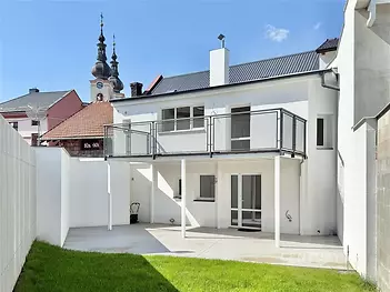 Přízemí domu s moderní fasádou, prostornou terasou a upraveným dvorkem. Dům má velká okna a nachází se ve zrekonstruované oblasti blízko kostela.