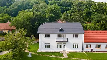 Velký rodinný dům s novou střešní krytinou a bílou fasádou, obklopený zelení. Klidná lokalita s velkou zahradou, vhodná k relaxaci.