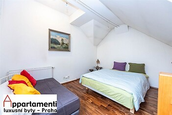 Podkrovní ložnice s dřevěnou parketovou podlahou, velkou postelí a světlými stěnami, nabízí útulný a moderní prostor pro relaxaci.
