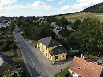 Žlutý rodinný dům s kvádrovým designem a šikmou střechou, umístěný u hlavní silnice v klidném zeleném prostředí příměstské oblasti.