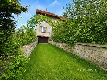 Zahrada s upraveným trávníkem a kamennou zdí u rodinného domu. Dům má moderní fasádu, červenou střechu a je obklopen zelení.