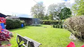 Zahrada s udržovaným trávníkem, dřevěnou lavičkou a trampolínou, obklopená zelení a keři, ideální pro rodinné aktivity nebo relaxaci