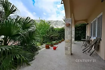 Zahradní terasa s dlažbou, obklopená bujnou zelení, ideální pro relaxaci. Kamenné detaily a přenosné židle vytvářejí příjemnou atmosféru.