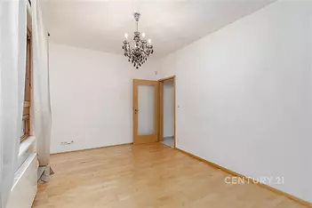 Světlý obývací pokoj s parketovou podlahou a moderním lustrem, bílými stěnami a velkými okny pro přirozené osvětlení.