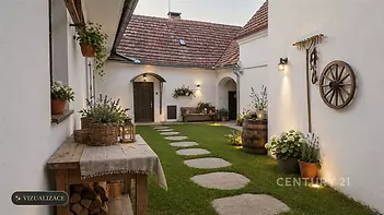 Venkovní dvorek s kamennou dlažbou, světlá fasáda, dekorativní lampy a květiny, rustikální styl, příjemná atmosféra pro relaxaci.