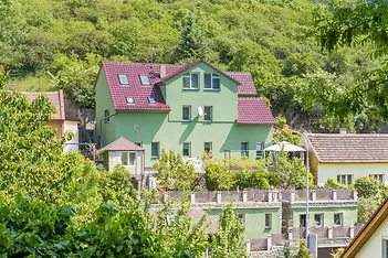 Dvoupatrový rodinný dům se zelenou fasádou, červenou střešní krytinou a upravenou terasovitou zahradou na svahu.