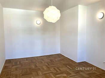 Prázdný pokoj s parketovou podlahou a bílými stěnami, moderní stropní svítidlo, dobrý stav, připravený k zařizování.