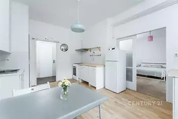 Světlá kuchyň s parketovou podlahou, moderním nábytkem a bílými spotřebiči, vedoucí do prostorné ložnice s velkými okny.