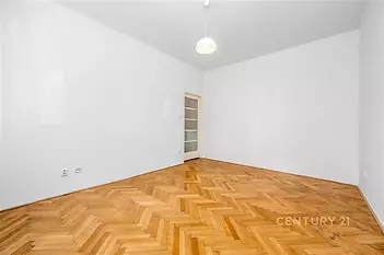 Prázdný pokoj s parketovou podlahou, bílými stěnami a moderním osvětlením. Vstupní dveře s prosklením zajišťují světlo do prostoru.