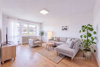 Světlý obývací pokoj, moderní styl, parketová podlaha, velká okna, pohodlná rohová sedačka, dekorativní rostliny, dřevěný nábytek.