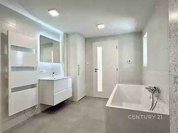 Moderní koupelna s dlažbou, volně stojící vanou a vestavěnými skříněmi, světlé provedení s jednoduchým osvětlením.