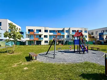 Dětské hřiště před moderním bytovým komplexem, obklopené zelení a lavičkami, nabízí klidné prostředí pro rodinné bydlení.