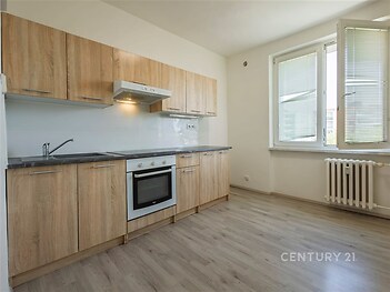 Moderní kuchyň s dřevěnými skříňkami, světlé parketové podlahy a velká okna zajišťující dostatek přirozeného světla.