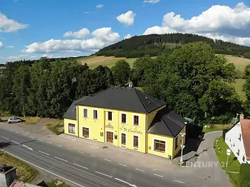 Žlutá budova s prostornou okolní krajinou, nachází se u hlavní silnice. Okolí tvoří les a pole, moderní stavba s okny v přírodním prostředí.