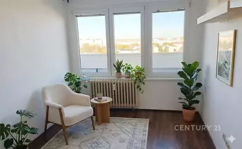 Světlý obývací pokoj s tmavou parketovou podlahou, velkými okny a moderním designem obsahuje pohodlné křeslo a pokojové rostliny.