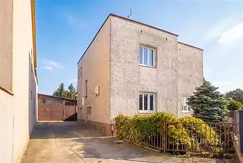Exteriér rodinného domu s velkou příjezdovou cestou, betonová fasáda, moderní okna a udržovaná zahrada okolo plotu.