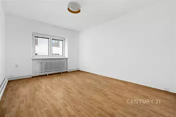Světlý pokoj s parketovou podlahou, bílými stěnami a velkým oknem, který poskytuje dostatek přirozeného světla, ideální pro moderní bydlení.