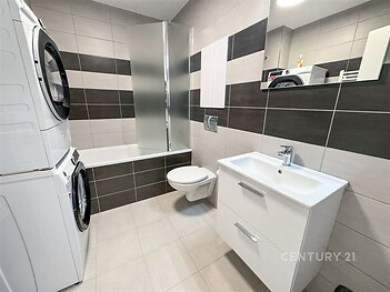 Moderní koupelna s dlažbou, vestavěným umyvadlem a úložnou skříňkou. Kombinace světlých a tmavých obkladů, pračka se sušičkou.